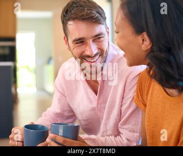 Loving Multi-Racial Couple Sitting On Sofa At Home With Hot Drink Laughing And Talking Stock Photo