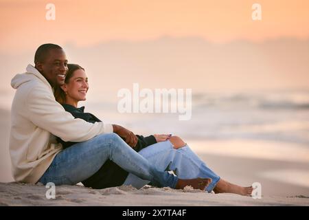 Casually Dressed Loving Young Couple Sitting On Beach Shoreline Hugging At Sunrise Stock Photo