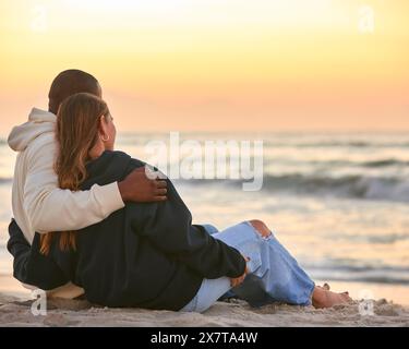 Rear View Of Casually Dressed Loving Young Couple Sitting On Beach Shoreline Hugging At Sunrise Stock Photo