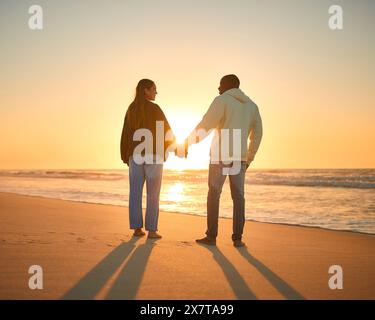Rear View Of Casually Dressed Loving Young Couple Holding Hands On Beach Shoreline At Sunrise Stock Photo
