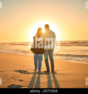 Rear View Of Casually Dressed Loving Young Couple Standing On Beach Shoreline Hugging At Sunrise Stock Photo