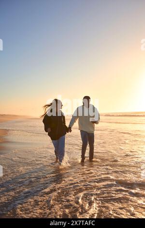 Casually Dressed Loving Young Couple Running Along Beach Shoreline Through Waves At Sunrise Stock Photo