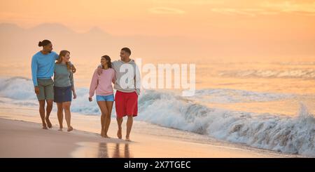 Hugging Couple With Friends In Casual Clothing On Vacation Walking Along Beach Shoreline At Dawn Stock Photo