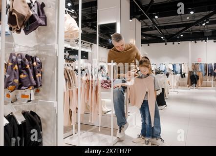 A father helps his young daughter pick out clothes in a brightly lit, modern retail store. Stock Photo
