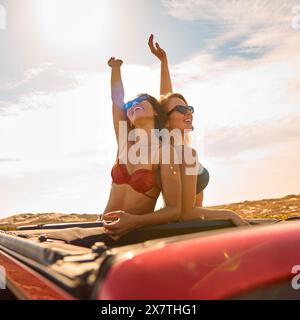 Two Women On Vacation Driving In Car On Road Trip Adventure To Beach Standing Through Sun Roof Stock Photo