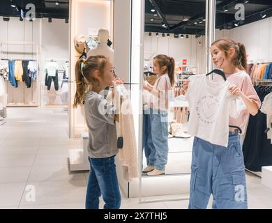 Two young girls, standing in a clothing store, are browsing through clothes on display with keen interest. Stock Photo