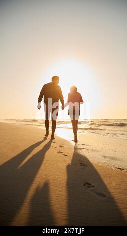 Rear View Of Retired Senior Couple On Vacation Walking On Beach Shoreline Holding Hands At Sunrise Stock Photo