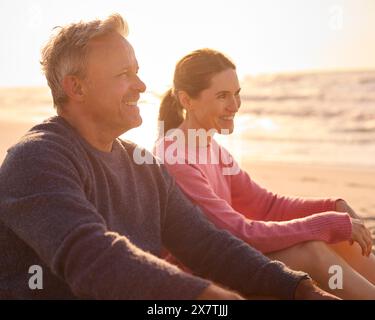 Retired Senior Couple On Vacation Sitting On Beach Shoreline Watching Sunrise Together Stock Photo