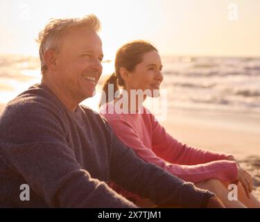 Retired Senior Couple On Vacation Sitting On Beach Shoreline Watching Sunrise Together Stock Photo