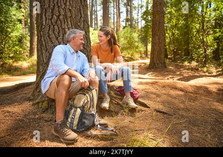 Retired Senior Couple Taking A Break Sitting At Base Of Tree On Hike Through Countryside Stock Photo
