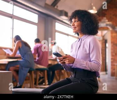 Smiling Businesswoman With Mobile Phone Working In Open Plan Office With Colleagues In Background Stock Photo