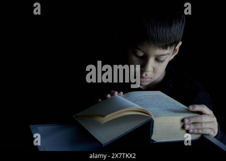 Latino child concentrating on reading a large book with a black background. Concept of reading in childhood Stock Photo