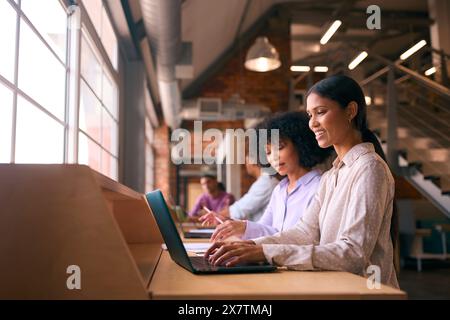 Two Businesswomen With Laptop Meeting In Office Discussing Documents With Colleagues In Background Stock Photo