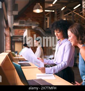 Two Businesswomen With Laptop Meeting In Office Discussing Documents With Colleagues In Background Stock Photo