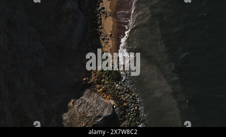 Aerial view of a rocky coastline with waves crashing against the shore. The image shows a mix of dark rocks, sandy beach, and ocean water. Stock Photo