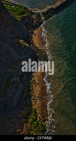 Aerial view of a rocky coastline with waves crashing onto the shore. The image shows a steep cliff on the left, a narrow sandy beach, and a breakwater Stock Photo