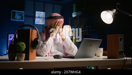 Tired arab businessman in traditional attire suffering migraine working at home office desk. Stressed Middle Eastern worker in living room answering emails getting headache from approaching deadline Stock Photo