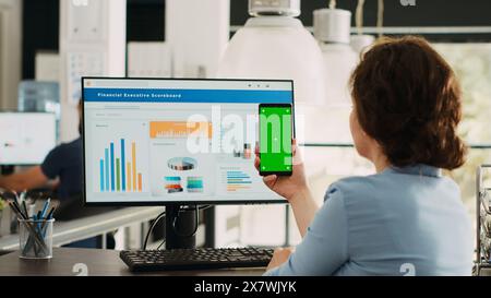 Employee looks at greenscreen on phone, working on business operations at desk. Person holding smartphone device with isolated display showing mockup template, blank layout screen. Stock Photo