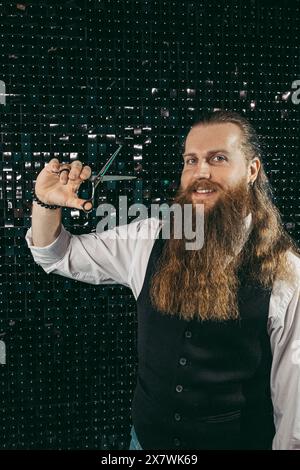 Bearded barber portrait with scissors .Man young barber hairdresser professional with long beard and hair in vest with scissors in hands portrait, cutting with scissors on black background. profession barber Stock Photo