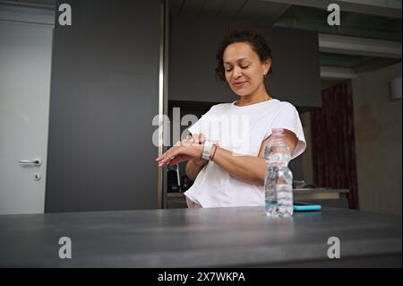 Multi ethnic smiling heathy young woman 40s, checking smart wrist watch. A bottle of pure water on the foreground. Fitness app, mobile app. People. He Stock Photo