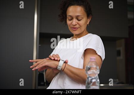 Beautiful Latin American smiling young woman 40s, in white t-shirt, checking her wearable smart wrist watch, standing at kitchen counter in the mornin Stock Photo