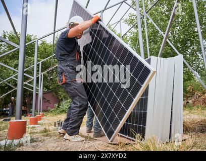 Workers carrying solar panels for installing in field. Technicians wearing workwear and helmets. Photo-voltaic collection of modules as a PV panel. Array as a system of photo-voltaic panels. Stock Photo