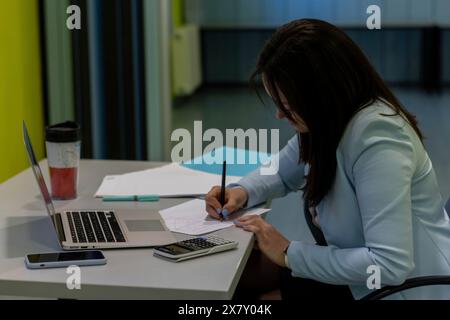 Side view of a confident businesswoman writing with pencil on notepad in the office with laptop and calculator on table Stock Photo