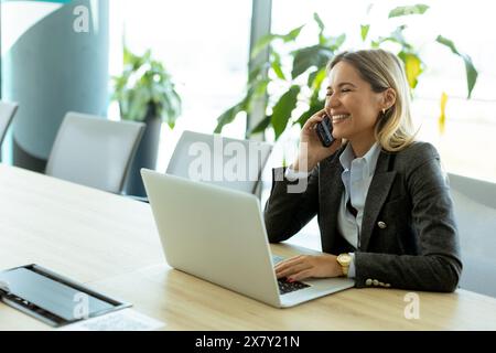 A professional smiling woman in a blazer engages in a phone call while working on her laptop in a well-lit office surrounded by plants. Stock Photo