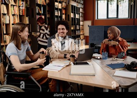 Group of diverse university students holding coffee cups sitting at table in library enjoying working on school project together, copy space Stock Photo