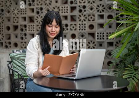 A young, positive Asian woman is reading a book at an outdoor table of a cafe, working remotely. A college student studying online. Stock Photo
