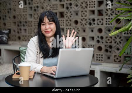 A young, positive Asian woman is smiling and waving her hand at the camera while sitting at an outdoor table with her laptop, working remotely from a Stock Photo