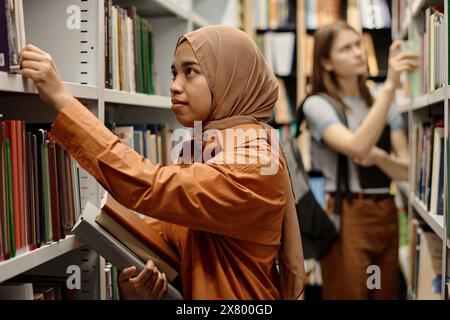 Selective focus shot of biracial female students choosing and taking books from bookshelves in college library Stock Photo