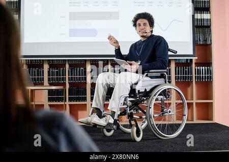 Over-the-shoulder shot of Middle Eastern student in wheelchair holding digital tablet speaking at conference in university library Stock Photo