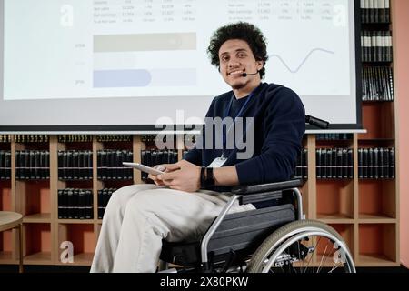 Portrait of cheerful young man with disability in wheelchair holding digital tablet smiling at camera while doing presentation Stock Photo