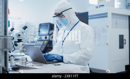 Electronic Manufacturing Factory: Engineer in Sterile Coverall Working on Laptop Computer, Examining a Circuit Board with Microchips and Testing New Electrical Equipment. Stock Photo