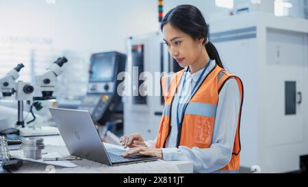 Portrait of an Asian Female Engineer in Orange Safety Vest Working on Laptop Computer at Electronic Manufacturing Factory. Technician Working on Daily Tasks and Research and Development Data. Stock Photo