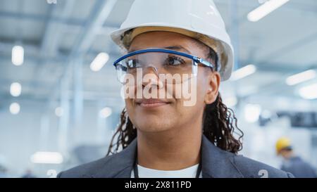 Close Up Portrait of a Happy and Smiling Middle Aged African Female Engineer in White Hard Hat Standing at Electronics Manufacturing Factory. Black Heavy Industry Specialist Supervising Work. Stock Photo