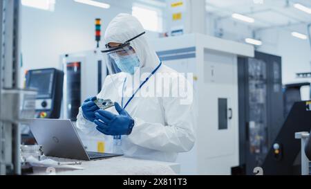 Electronic Manufacturing Factory: Engineer in Sterile Coverall Working on Laptop Computer, Examining a Circuit Board with Microchips and Testing New Electrical Equipment. Stock Photo