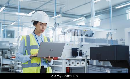 A Focused and Professional Asian Female Engineer in White Hard Hat Standing with Laptop Computer at Electronic Manufacturing Factory. Successful Employee Working on Daily Tasks Stock Photo