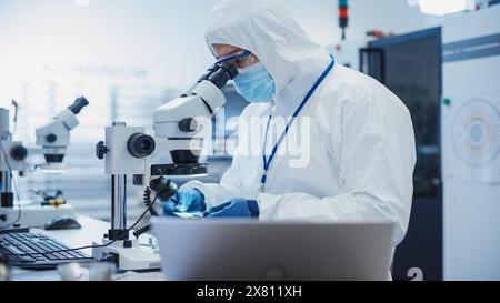 Electronic Manufacturing Factory: Engineer in Sterile Coverall Examining a Circuit Board with Microchips Through a Microscope. Specialist Comparing Results on Laptop Computer Stock Photo