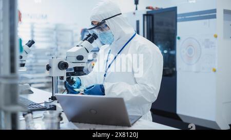Portrait of an Engineer in White Safety Coverall Working on Laptop Computer at Electronic Manufacturing Factory. Technician Working on Daily Tasks and Inserting Analytic Science Data. Stock Photo