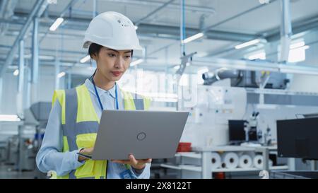 A Focused and Professional Asian Female Engineer in White Hard Hat Standing with Laptop Computer at Electronic Manufacturing Factory. Successful Employee Working on Daily Tasks Stock Photo