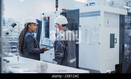 Two Diverse Professional Heavy Industry Engineers Wearing Safety Uniform and Hard Hats Working on Laptop Computer. African American Technician and Worker Talking Next to a Factory Machine. Stock Photo