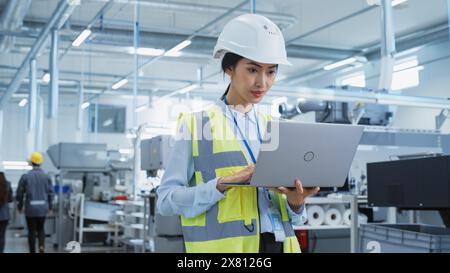 A Focused and Professional Asian Female Engineer in White Hard Hat Standing with Laptop Computer at Electronic Manufacturing Factory. Successful Employee Working on Daily Tasks Stock Photo