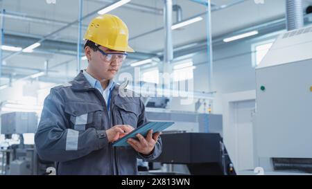 Portrait of a Young Asian Man, Working as an Engineer at a Factory Facility, Wearing Work Jacket and a Yellow Hard Hat. Heavy Industry Specialist Working on Tablet Computer. Stock Photo