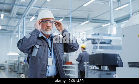 Close Up Portrait of a Middle Aged, Successful Male Engineer in White Hard Hat and Safety Glasses, Standing at Electronics Manufacturing Factory. Heavy Industry Professional Posing for Camera. Stock Photo