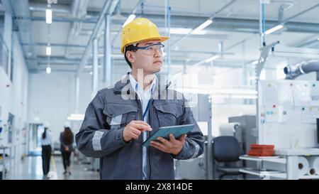 Portrait of a Young Asian Man, Working as an Engineer at a Industrial Facility, Wearing Work Jacket and a Yellow Hard Hat. Heavy Industry Specialist Walking and Using Tablet Computer. Stock Photo