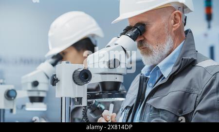 Middle Aged Industrial Science Engineer Working at a Desk in a Electronics Factory Facility, Using Microscopes to Work on Small Manufacturing Production Parts and Details. Stock Photo
