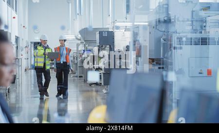 Two Professional Heavy Industry Employees Wearing Hard Hats at Factory. Checking and Discussing Industrial Machine Part, Working on Laptop Computer. Asian Engineer and Middle Aged Technician at Work. Stock Photo