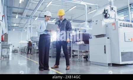 Two Young Asian Heavy Industry Engineers Walking with Their Backs to Camera, Using Laptop Computer, Discussing Production of a New Component. Two Employees at Research and Development Factory. Stock Photo